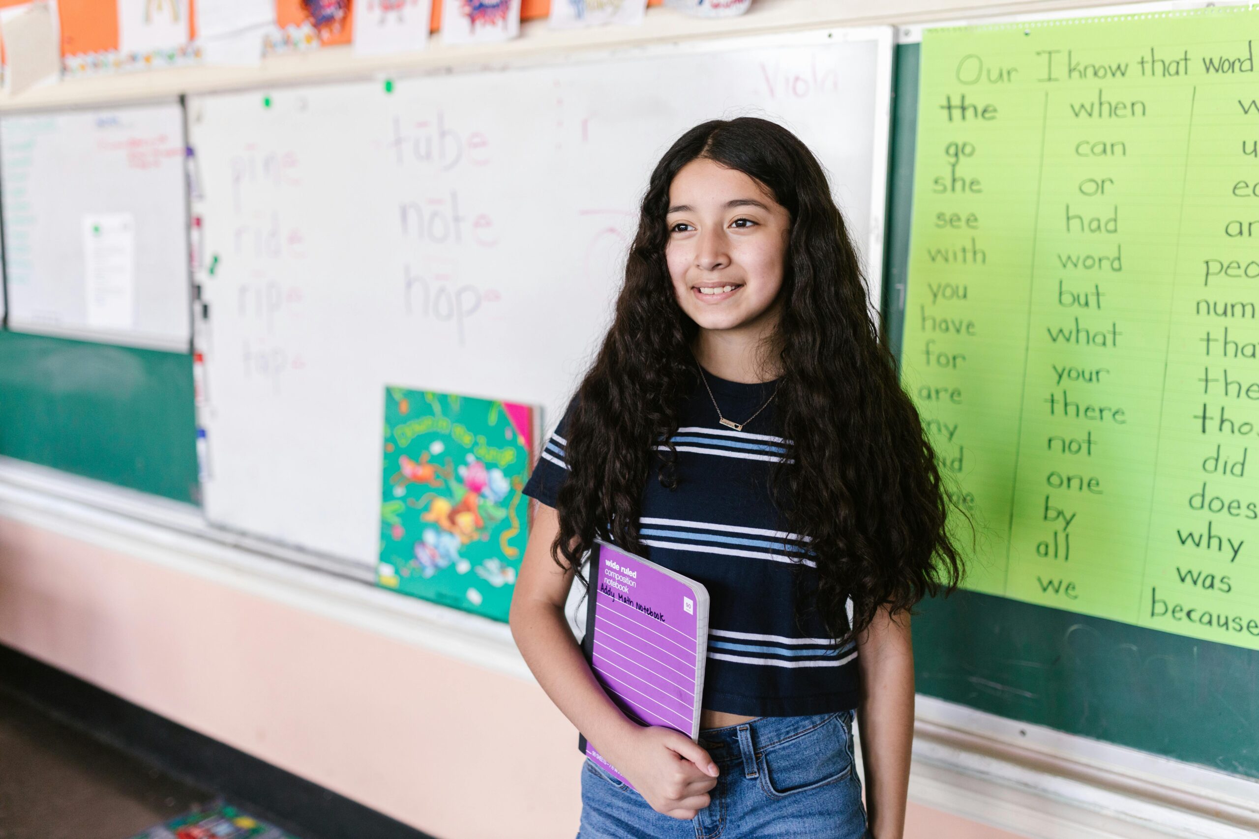 Young girl in a classroom holding a notebook, smiling at the camera with a backdrop of educational materials.